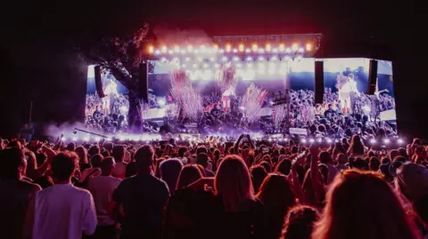 Crowds attending Olivia Rodrigo's headliner set at BST Hyde Park in 2025. The evening audience is being lit up by an intricate light show on the stage.