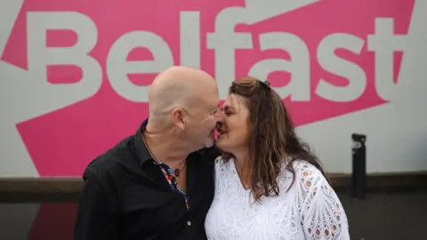 PA Media A man and woman kiss in front of a pink and white Belfast sign.