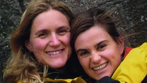 Two sisters with smiling faces hugging. Kim Leadbeater is to the left with blonde hair with Jo Cox who has brown hair. 
