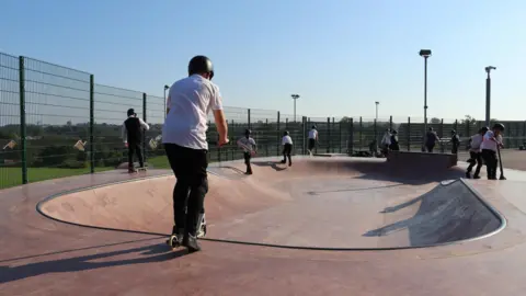 Turner Schools Young children in school uniform ride skateboards and scooters in a skatepark.