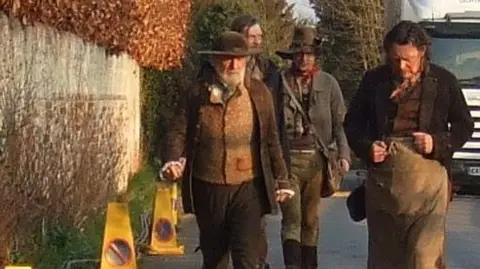 Margaret Blake Four actors of Les Miserables walking down the street in Ewelme, wearing their film clothes and makeup. There is a white vehicle behind them and several yellow cones by a wall they are walking by.