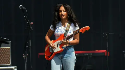 Getty Images Lily Fontaine, the singer with English Teacher. She is wearing a white T-shirt and jeans, and playing a red guitar. 