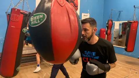 Coach Paul McIlvenny has short dark hair and a beard. He is striking a red and black punchbag hanging from the ceiling of the gym, which has blue walls and a light-coloured wooden floor. Paul is wearing a black "SAVE OUR CLUB" t-shirt.