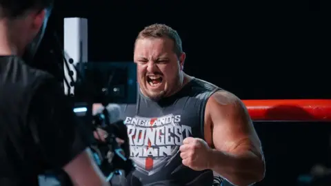 Ben Musgrove Dean Mason - a man with broad shoulders and muscles - has his mouth open wide and is frowning whilst clenching his first. He is wearing a grey shirt that says England's Strongest Man and a cameraman is standing in front of him.