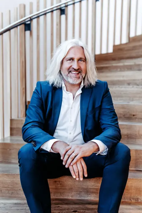 Christian Ege Head shot of Christian Ege with shoulder-length grey hair and beard sitting on some wooden steps. He is wearing a blue suit with a white shirt with the top buttons unopened.