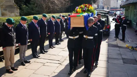 Royal Marine servicemen wearing dark suits with white belts, carrying the wooden coffin of Jim Wren. The coffin is covered with a flag and a large bouquet of spring flowers. Beside the paved walkway stands a long row of former and currently serving marines, wearing green caps and standing with their heads bowed as the coffin passes.