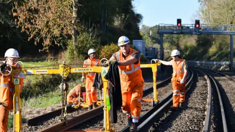 Men in orange hi-vis suits walk down train tracks with a yellow piece of machinery