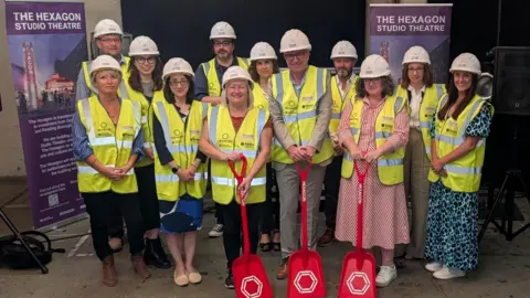 A group of people wearing smart work clothing, with high visibility vests and white hard hats. The three people at the front are holding red shovels. They're all smiling at the camera with banners either side of them about the new Studio Theatre at the Hexagon.