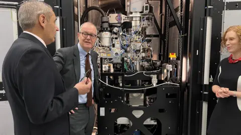 Martin Giles/BBC Patrick Vallance listening to a man speaking. A woman is standing to the side of a large bit of machinery with the two men on the other side of it. 