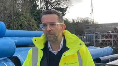 A man with glasses wearing a neon yellow jacket standing in front on blue piping