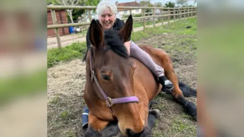 Kelly Allen, who has grey hair and is wearing a black top and grey tracksuit bottoms, smiles as she sits on top of DD, who is lying down. DD is a brown mare and is on the grass of her paddock, with a fence behind her.