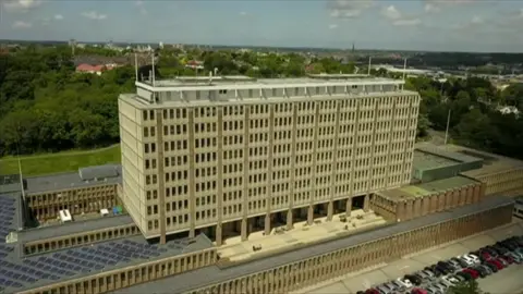 Shaun Whitmore/BBC Norfolk County Hall, captured in a drone image. The building was built in the 1970s in the Brutalist style. The building is a beige colour, with 11 full bays, flanked by two half bays, over ten floors in the main tower section, with a further four floors below in stepped levels. Cars can be seen parked at the front, and there are green lawns and trees in the background, with the city of Norwich partially visible beyond.