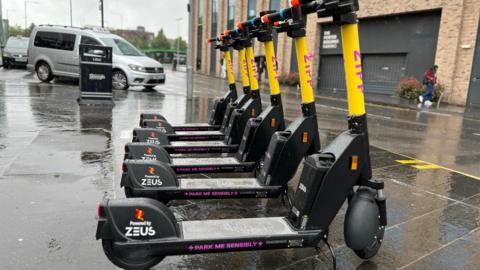 Six Zipp e-scooters outside Slough rail station. They have a yellow body, with Zipp written in pink on them. "Park me sensibly" is written on a footplate 