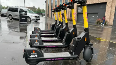 BBC Six Zipp e-scooters outside Slough rail station. They have a yellow body, with Zipp written in pink on them. "Park me sensibly" is written on a footplate 
