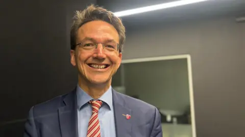 BBC Kirsten Morel is dressed in formal attire, wearing a navy blue suit jacket, a light blue shirt, and a red-and-white striped tie. The background features a dark wall with a bright horizontal light fixture and a reflective surface, suggesting an indoor professional or studio environment