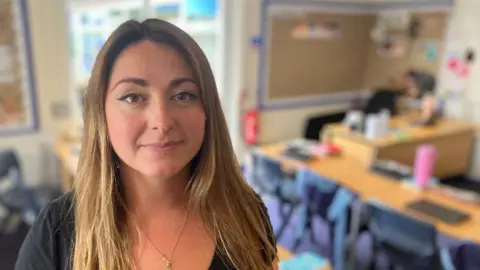 Mawnan Primary School assistant head teacher Catherine Prosser stands in a classroom. She is in her 30s, with long light-brown hair and wearing a black top.