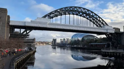 The Tyne Bridge, covered in scaffolding, sits above the River Tyne with the silver dome shape Glasshouse Centre for Music on the right. It's a cloudy day and buildings can be seen in the distance as the river bends to the left.