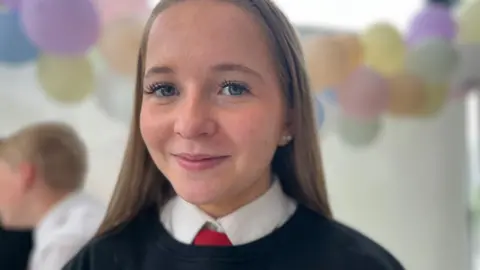 Ellie looks at the camera. She is wearing the school uniform of a black jumper, white shirt and red tie
