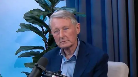 Sir David Normington, a grey haired man in a blue open neck shirt and jacket, sits in front of a microphone in the Newscast studio