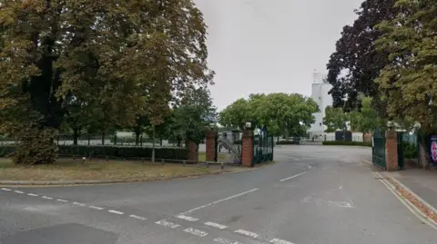 Google The entrance to Kempton Park racecourse seen in a Google Streetview image. There are large metal gates which are open and one of the stands can just be seen behind some trees.