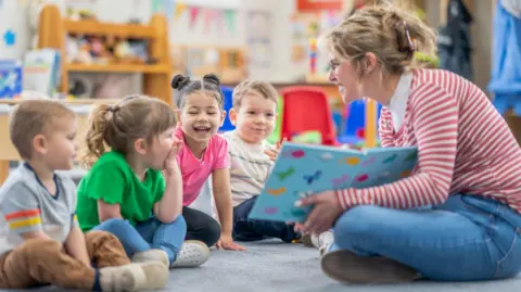 A woman reads a book to four young children in a nursery room. She is sat cross-legged on the floor with a blue book open to the two girls and two boys, who are smiling.