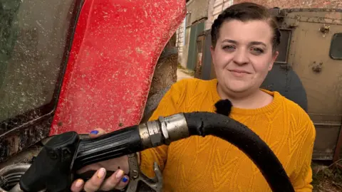 Harriet White is wearing a mustard-coloured jumper and standing next to a red tractor. She is filling the tractor up with diesel. She has short brown hair.