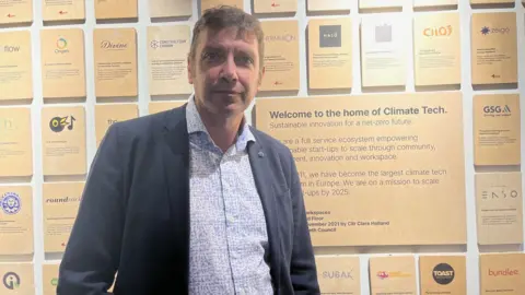 Andrew Wordsworth wearing a blue jacket over a blue and white patterned shirt with wooden blocks with writing on on a wall behind him