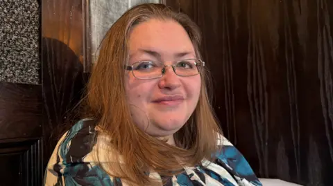 A woman with light brown shoulder-length hair wearing a floral blouse and silver-rimmed glasses looking into the camera. 