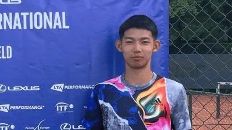 A young man with short dark hair and a multi-coloured sports top standing in front of a blue advertising screen with a clay tennis court partially visible behind him