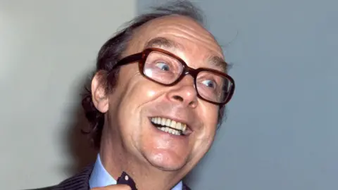 BBC Eric Morecambe looks to the side of the camera. He is smiling and straightening his tie. He wears brown, thick-framed glasses. He has short dark hair.