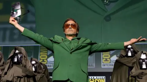 Robert Downey Jr holds the silver mask at the end of his outstretched arm