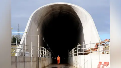 PA Media A worker in orange safety gear walks toward the entrance of a large concrete HS2 railway tunnel under construction, framed by scaffolding on both sides.