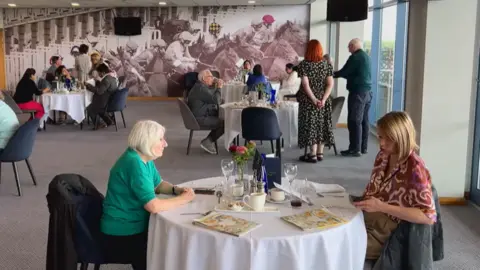 Two women in the foreground sit across from each other on a round table, with menus and glasses in the middle of it. In the background are three other round tables with people sitting around them, and the back wall is painted with a photo of people racing horses.