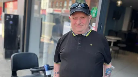 Jonny Humphries/BBC Jimmy Williams, wearing a black polo-neck t-shirt, black baseball cap and sunglasses on his head, smiles at the camera on the street at County Road.