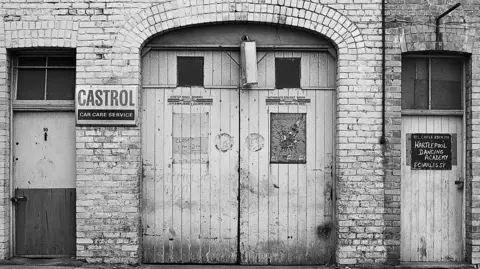Peter Benson A black and white image of closed garage doors between brick pillars. One one side is an advertisement for Castrol oil. On the other a sign in chalk on a smaller door suggests reads Hartlepool Dancing Academy.