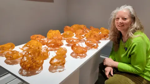 Helen Fox is crouched down with her sculptures. They are a collection of orange netting morphed into the shape of pebbles. 