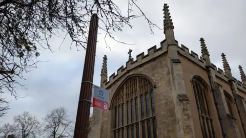 Alex Harris/BBC View of St John's Street in Cambridge showing a street light without a lantern at the top. There is a bus stop sign attached to the post and an ornate church in the background