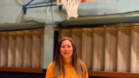 A long-haired woman in an orange top stands in front of a basketball net.