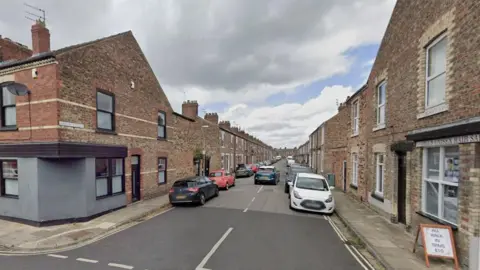 A street view image shows a residential area with two-storey homes running along on both sides. Parked cars are next to the curb. A sign standing on the pavement next to a hair salon on the right corner of the road reads "All walk in trims £10".