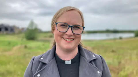 Reverend Beth Cope smiling at the camera. She is wearing a dog collar, glasses grey jacket. Behind him is a lake surrounded by grass, out of focus. 