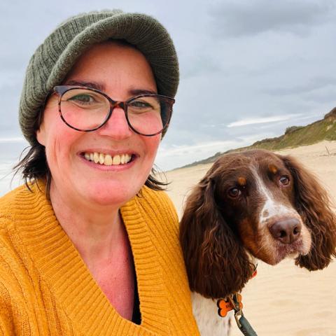 Sophie Leggett with glasses, a yellow jumper and a hat, is stood next to a dog on the beach