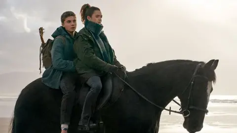 True Brit Entertainment A young woman rides a dark coloured horse. There is a teenage boy riding on the same horse behind her. They are riding on a beach. They wear coats and the boy has a rucksack on his back. 