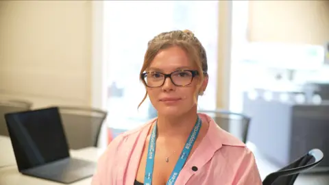 Jamie Niblock/BBC Alice is sitting on a chair in an office. There is a laptop and more chairs in the background. She is wearing a pink shirt and glasses and has a blue lanyard with white writing.