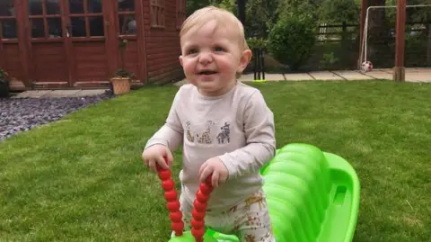 A toddler is sitting on a green plastic caterpillar toy, holding on to its red antennaes on grass in a garden with a brown shed, patio and football goalpost behind him. He is smiling and wearing a beige top and trousers with zoo animals on them.