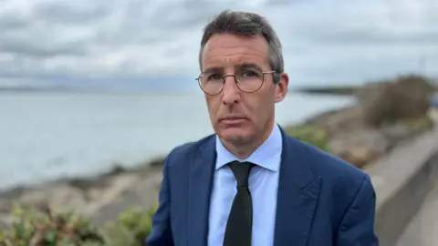 Andrew Muir, a man with grey hair and glasses, wearing a dark grey suit, white shirt and black tie.