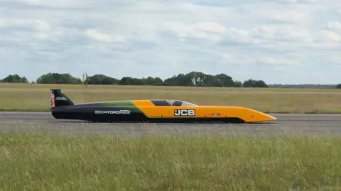 A photograph of the JCB vehicle on a runway. It is a long vehicle with a yellow front with "JCB" on it. The back half of the car is dark green and black with white writing. To the front and back is grass and it is on a grey surface.