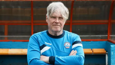 John Fairhall/BBC Steve Pitt is wearing a blue Braintree sweater in front of a dugout at the club's stadium. He has long grey hair and has his arms folded.