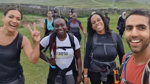 Four people are standing in a field taking a selfie. One woman on the left is holding up her hand in a peace sign. Two women are in the middle with backpacks and walking gear on. Sameed Asghar is on the right, wearing a red t-shirt and a rucksack.