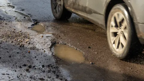 Getty Images A gritty pothole on a road, full of rainwater, with the wheel and side of a silver car visible next to it 