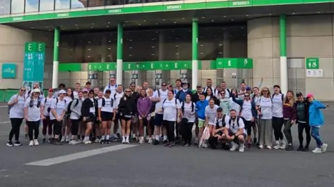 Wigmore Primary School A group of nearly 60 men and women standing outside Wembley Stadium's front gates. They are all wearing T-shirts, shorts or leggings and trainers or walking boots. They are all smiling and some are waving. 